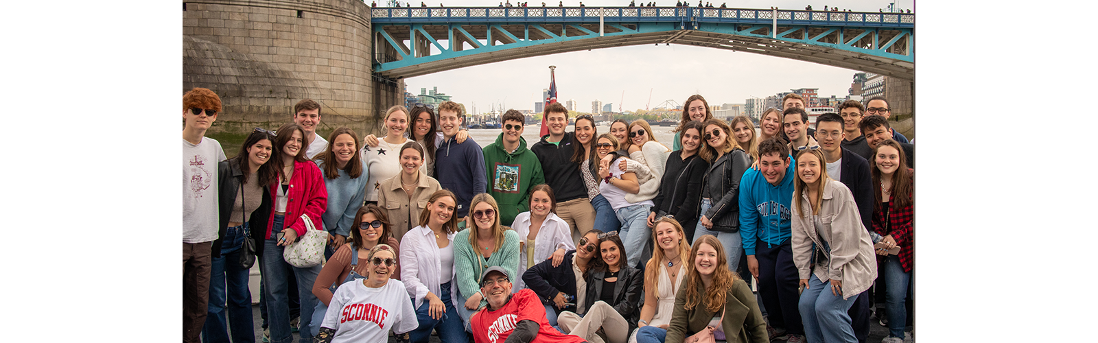 Students are gathered together under one of London's iconic bridges. The students are all smiling at the camera and hugging.